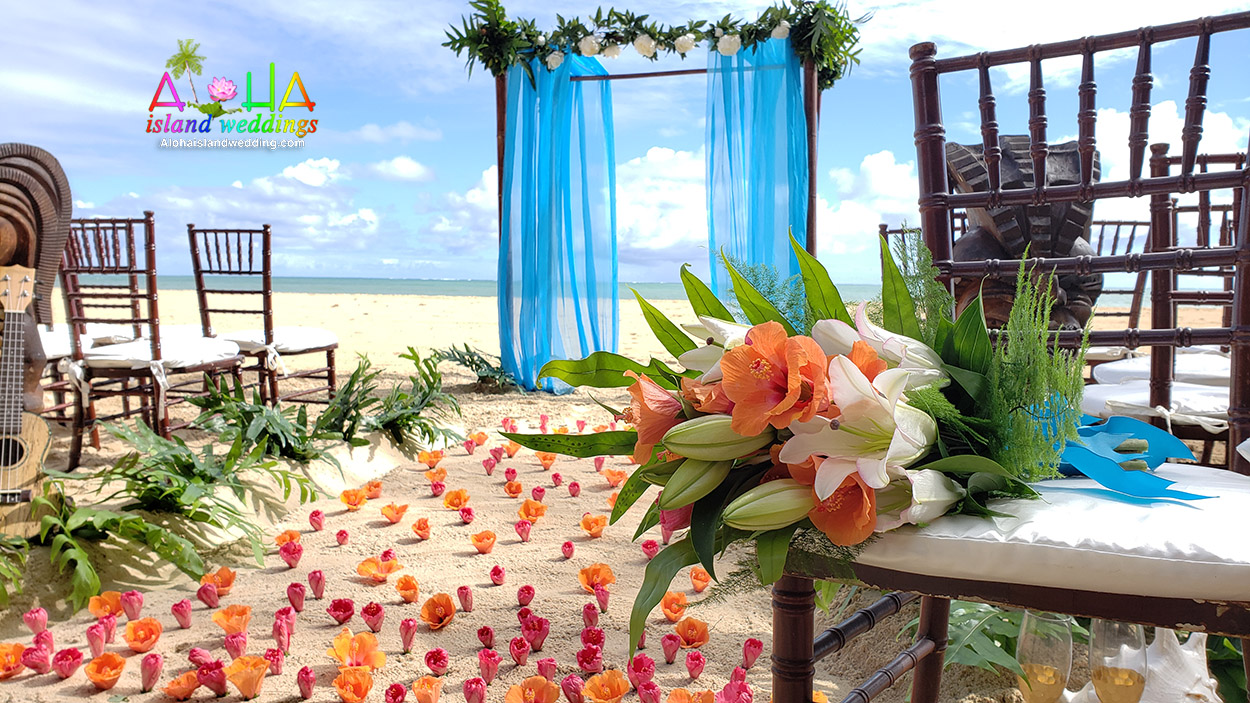 Beach setup with turquoise drapes, wooden chairs, coral petals and a tropical flower arrangement